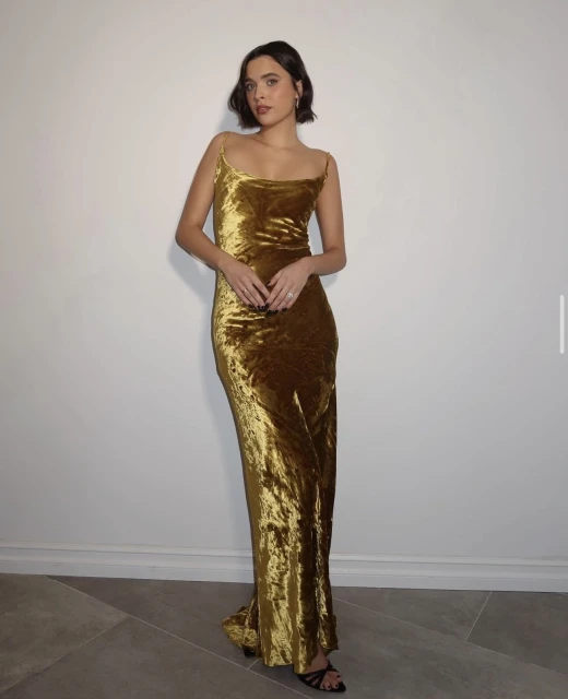 Portrait of a woman in a gold velvet dress. She is indoors with neutral lighting, posing against a white wall and grey stone floor.