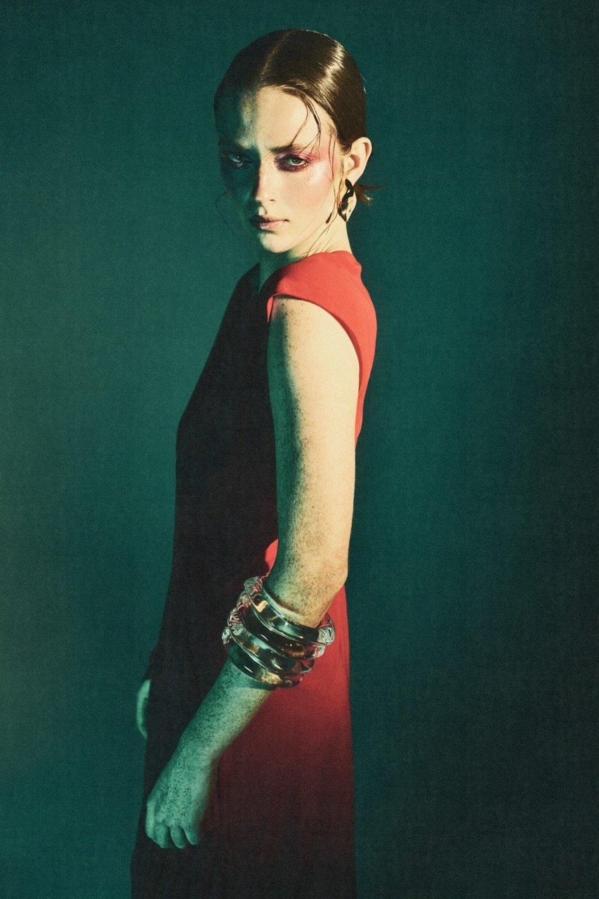 A woman in a red dress poses against a teal backdrop, wearing several metal bracelets, with dramatic pink makeup and slicked-back hair.