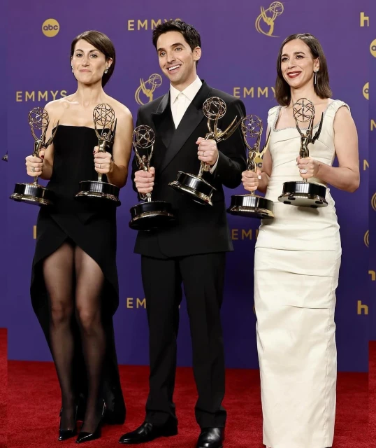 Three smiling people in formal attire, holding Emmy awards, stand in front of a purple backdrop at an entertainment event.