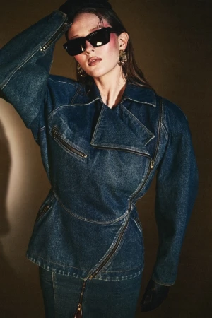 Brunette woman in a denim jacket and skirt with black sunglasses posing indoors in a fashion shot with a neutral background.