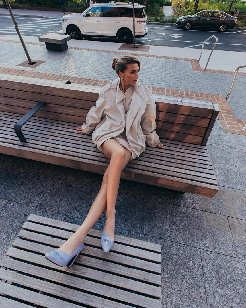 Elegant woman in trench coat and blue high heels sits on a bench in an urban environment, with cars visible in the background.