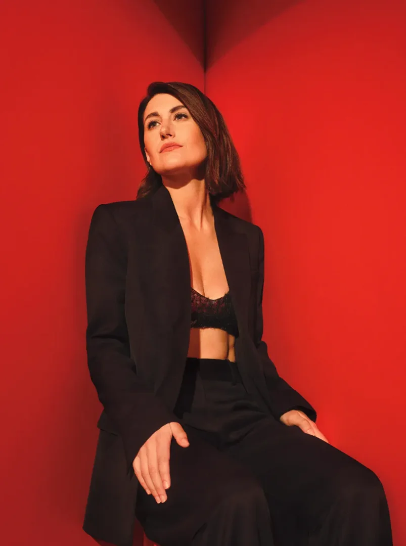 Elegant woman in a black pantsuit and lace bralette sits in front of a solid red wall, gazing upwards with a neutral expression.