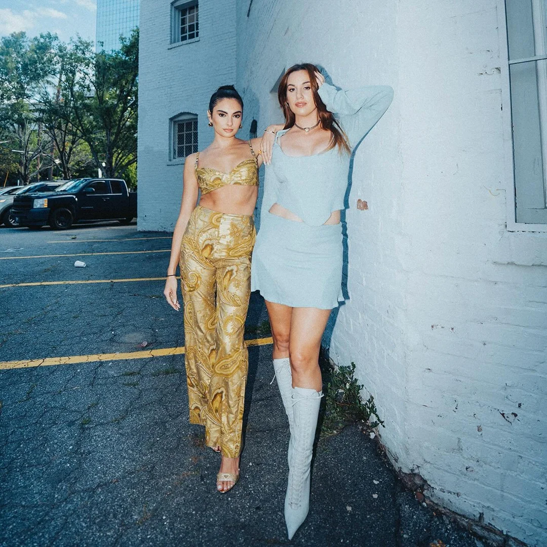 Two women pose in fashionable outfits on a city street next to a brick building, featuring contemporary style and urban backdrop.