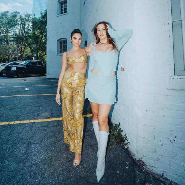 Two women pose in fashionable outfits on a city street next to a brick building, featuring contemporary style and urban backdrop.