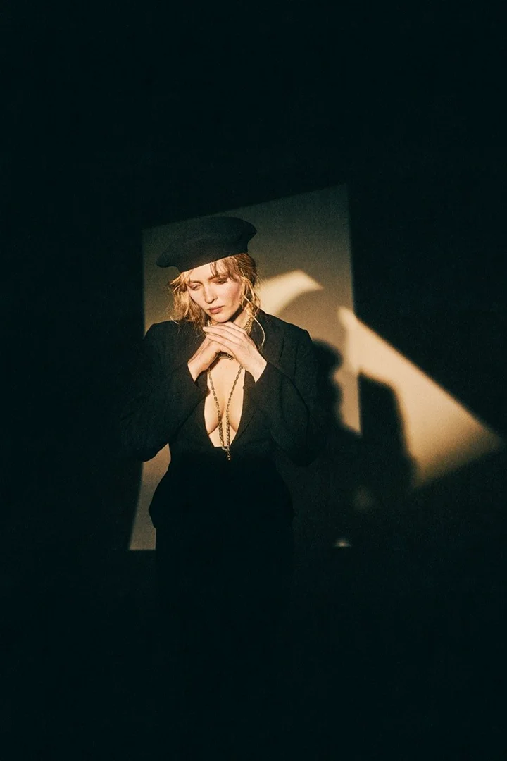 Woman in black suit and hat poses in a dark room illuminated by a soft, golden light highlighting her face and hands.