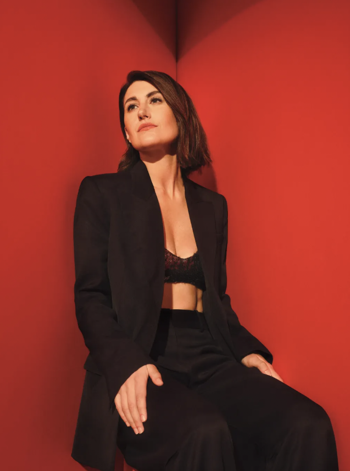 Portrait of an adult woman with dark hair wearing a black suit, posing in a red-walled photo studio setting. Looking upward.