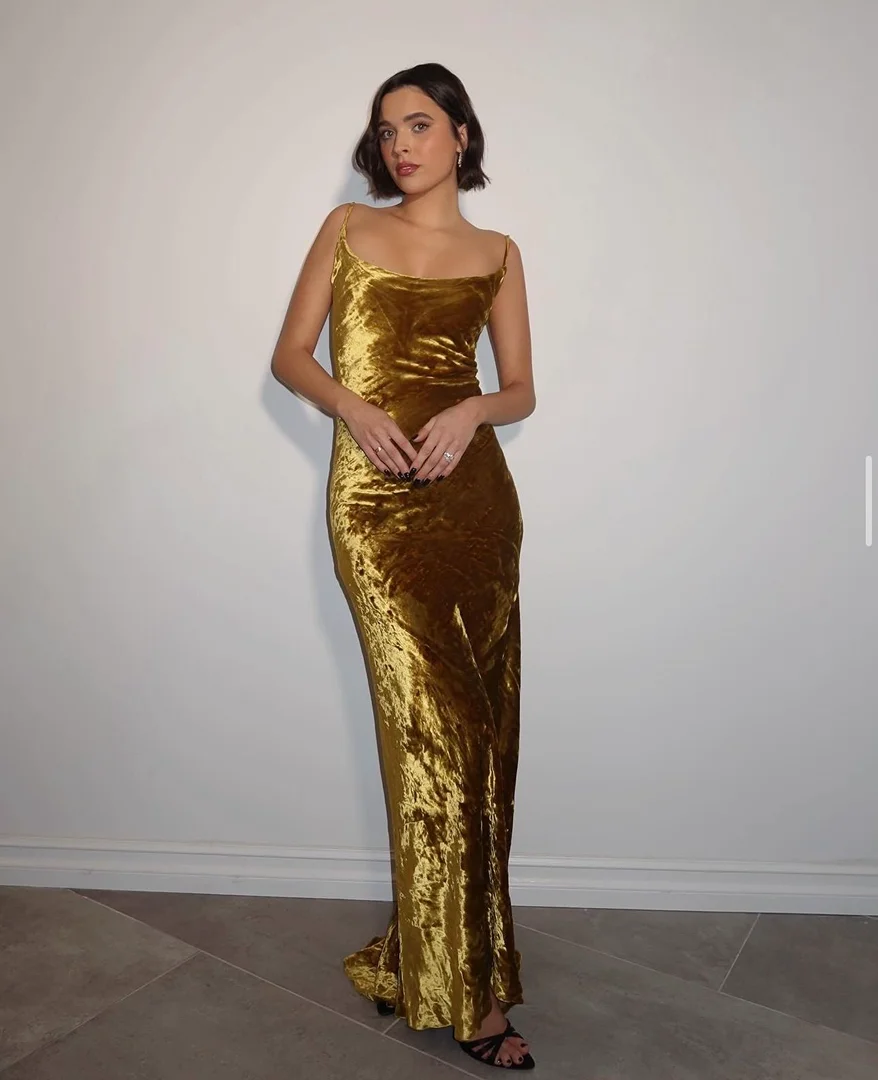 Adult woman in a gold-colored velvet dress posing against a white wall with gray flooring, looking directly at the camera with neutral expression.