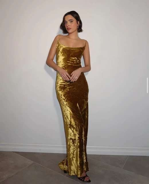 Adult woman in a gold-colored velvet dress posing against a white wall with gray flooring, looking directly at the camera with neutral expression.