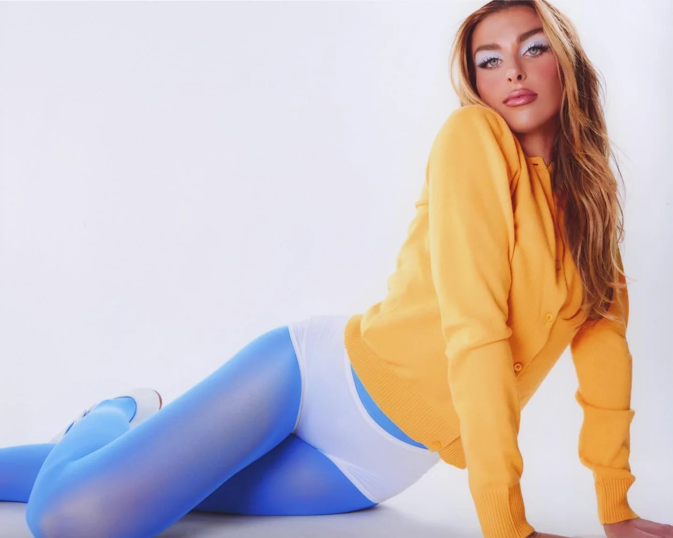 Attractive woman in a yellow cardigan and blue tights reclines against a white backdrop, striking a pose while looking at the camera.