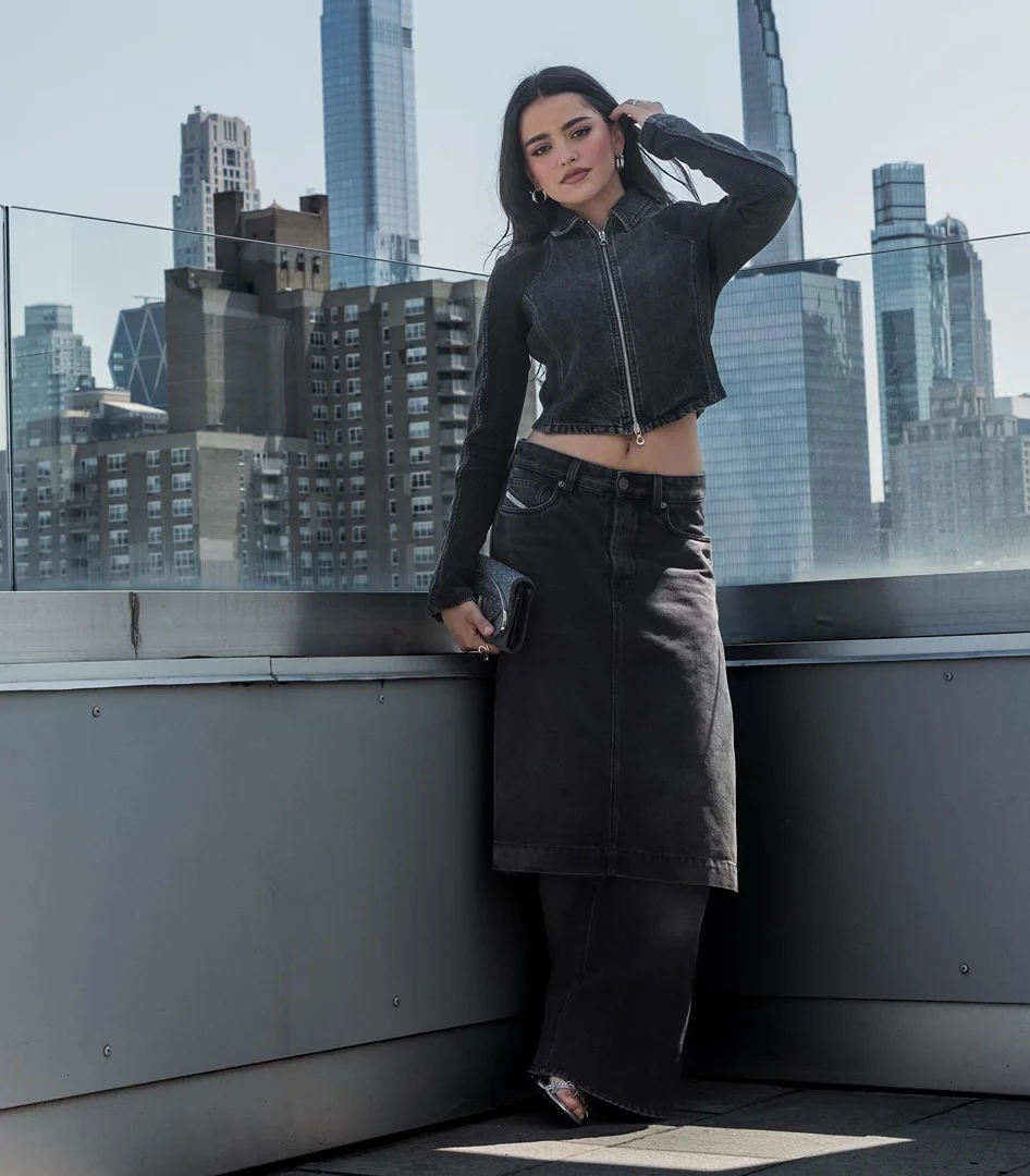 Woman poses in black denim jacket and long skirt on a rooftop with a city skyline backdrop under bright sunlight.