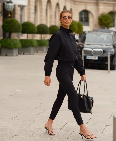 Woman in black athletic jacket and leggings walking on a city street, carrying a black handbag and wearing sunglasses.