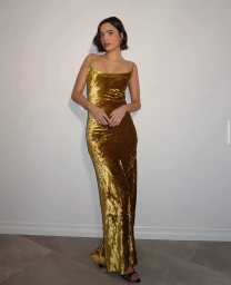 Woman with short dark hair wearing a long gold velvet dress standing against a white wall.