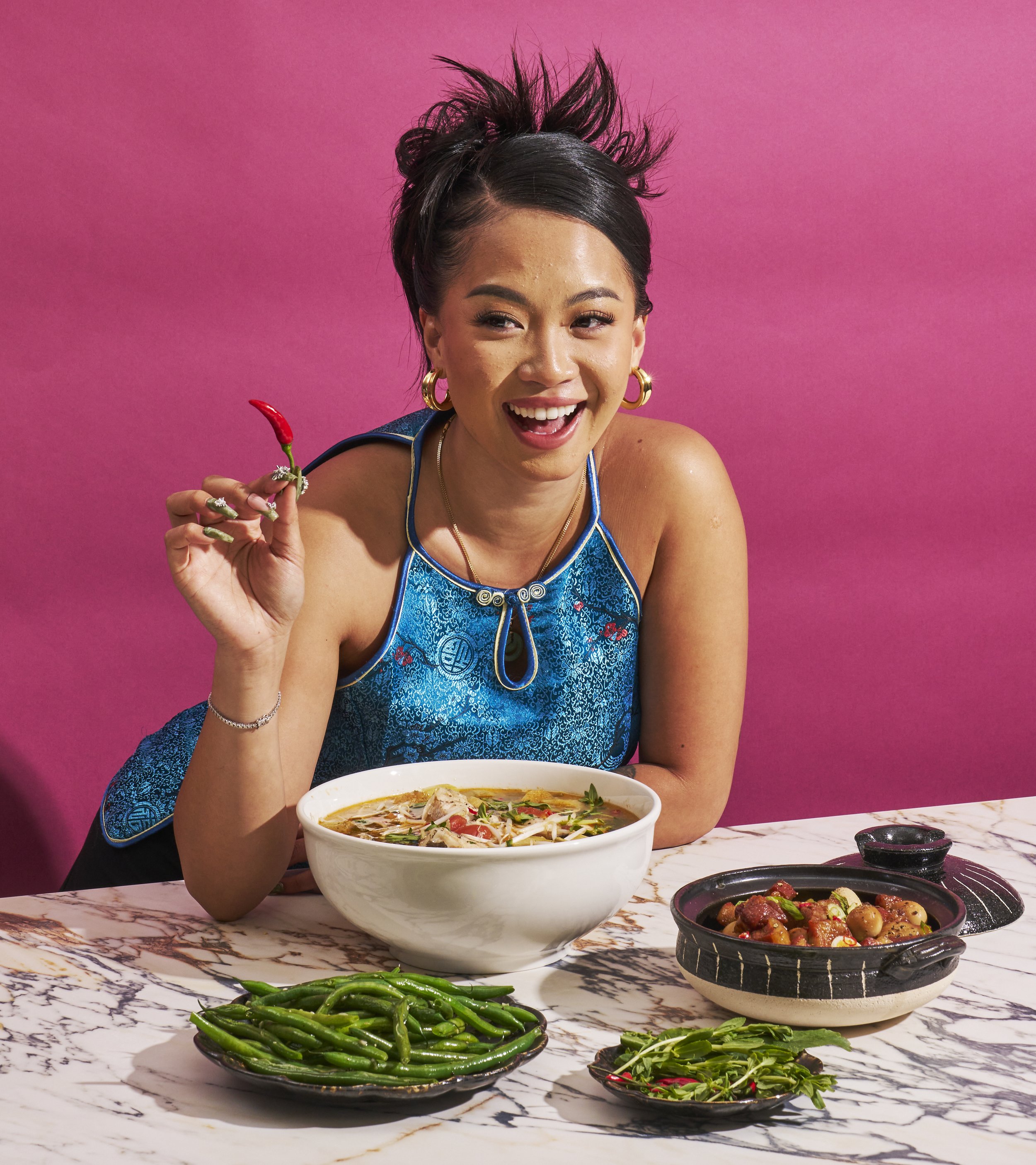 Woman smiles holding a chili pepper, seated with a bowl of pho, side dish of glazed meat, and green beans on a marble surface against pink.