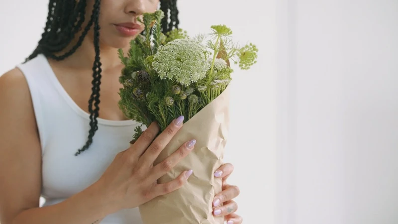 Young woman with box braids holds a bouquet of fresh flowers wrapped in brown paper against a white wall, indoor setting, close-up shot.