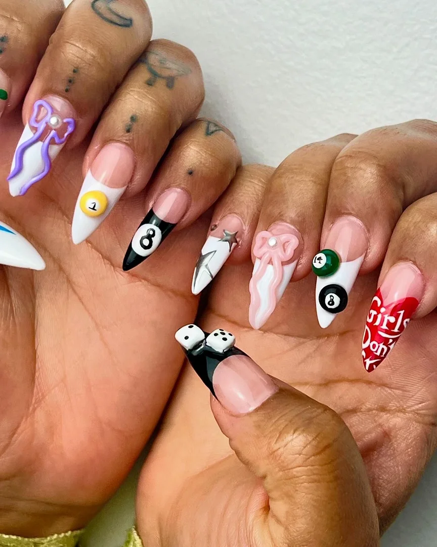 Close-up of a person's hands with long, decorated stiletto nails featuring nail art with dice, bows, pool balls, and writing.