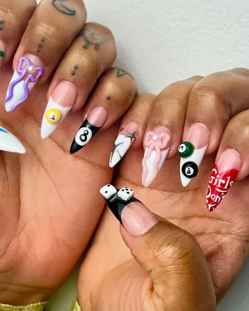 Close-up of a person's hands with long, decorated stiletto nails featuring nail art with dice, bows, pool balls, and writing.