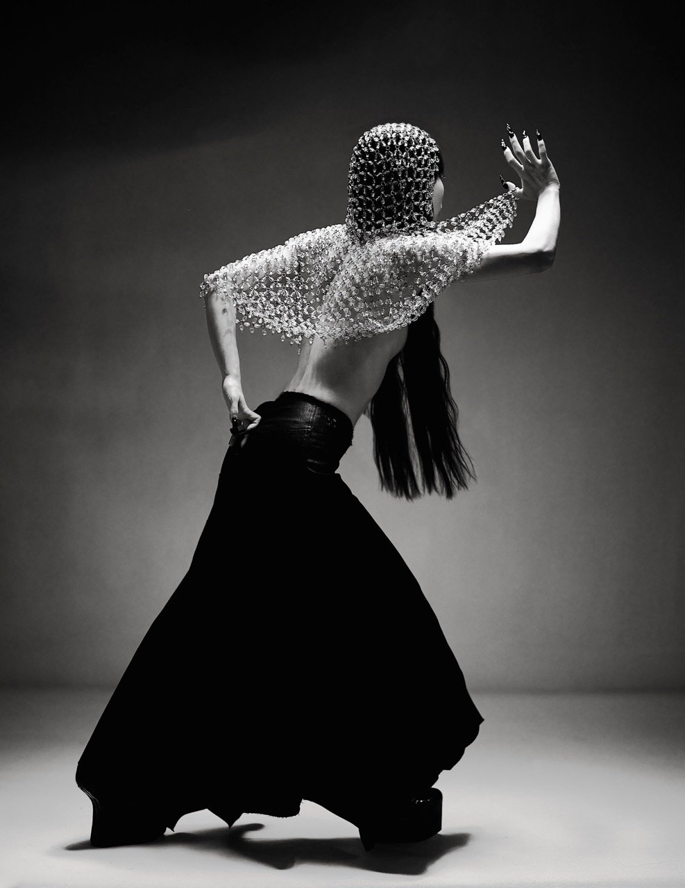 Monochrome studio shot of a woman in avant-garde fashion, wearing chainmail and a black skirt, showcasing movement and dramatic style.