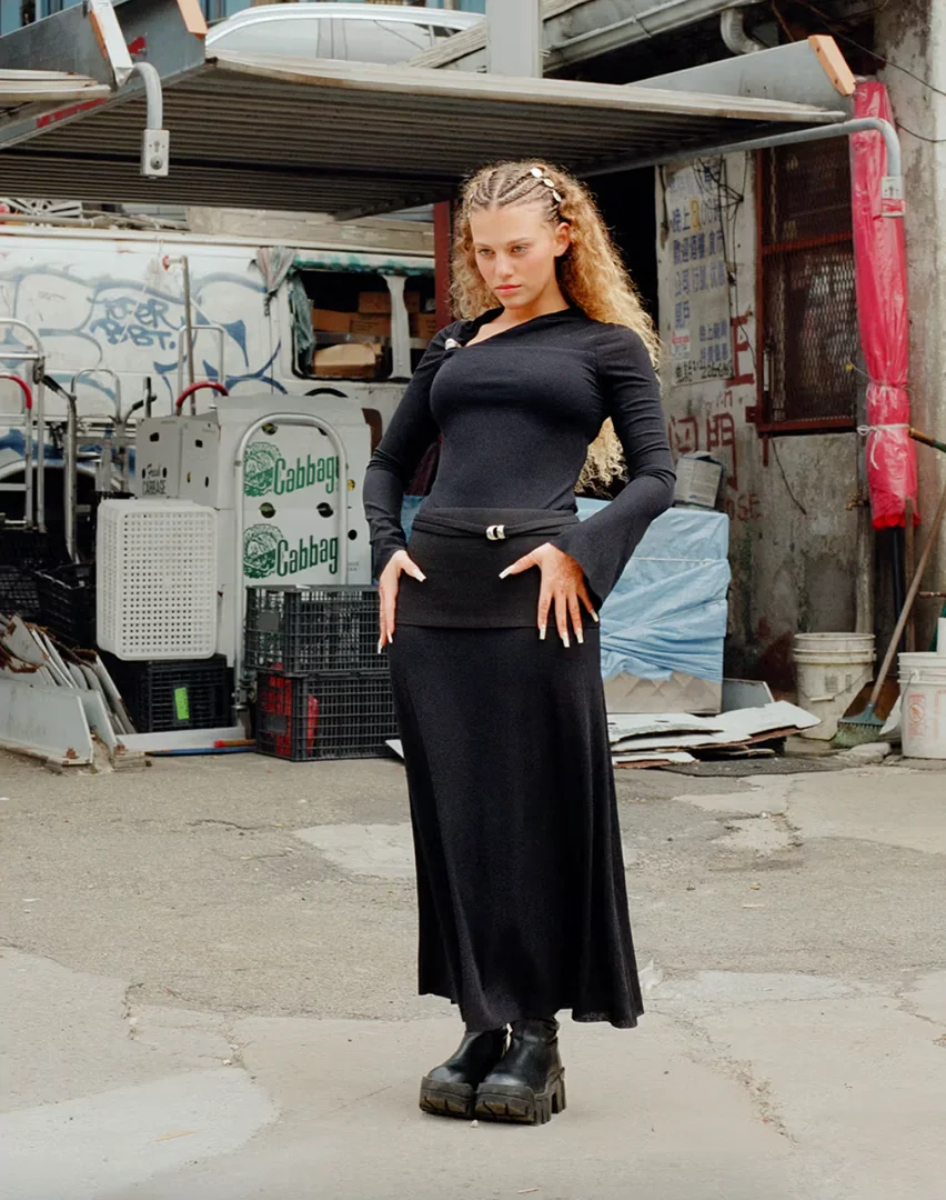 A woman with blonde braids poses in black dress with platform boots on a concrete street, with a white truck and building in the background.