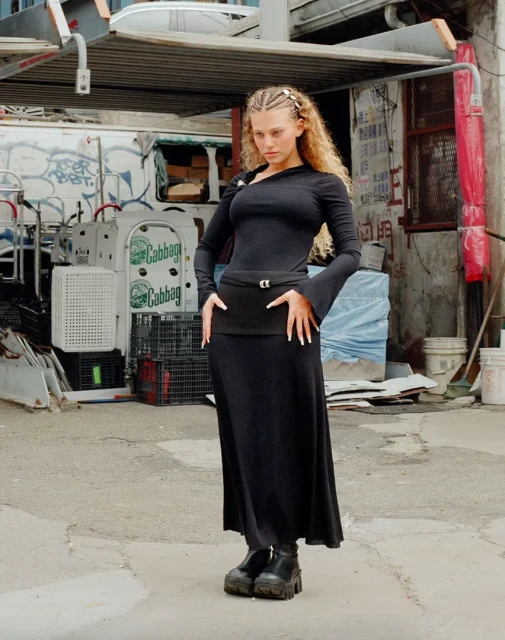 A woman with blonde braids poses in black dress with platform boots on a concrete street, with a white truck and building in the background.