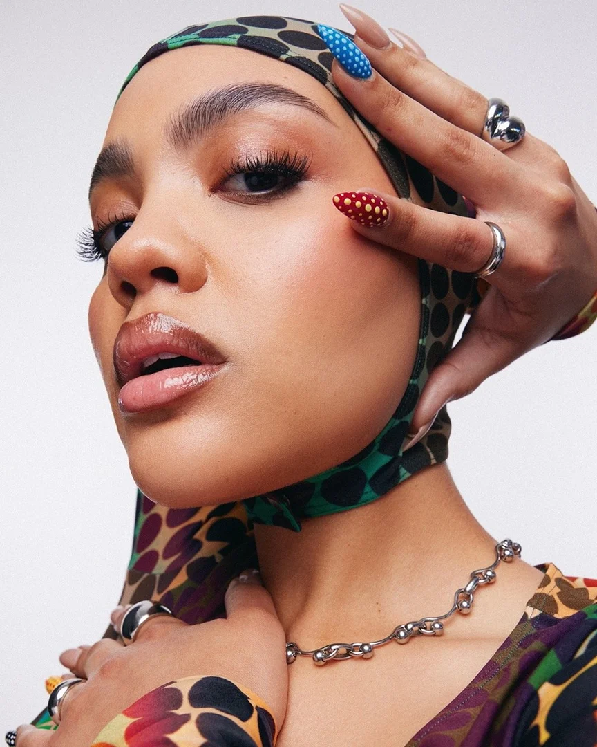 Close-up studio shot of a woman wearing a printed headscarf, silver necklace and rings, and multicolored polka dot nails, posing with hands.