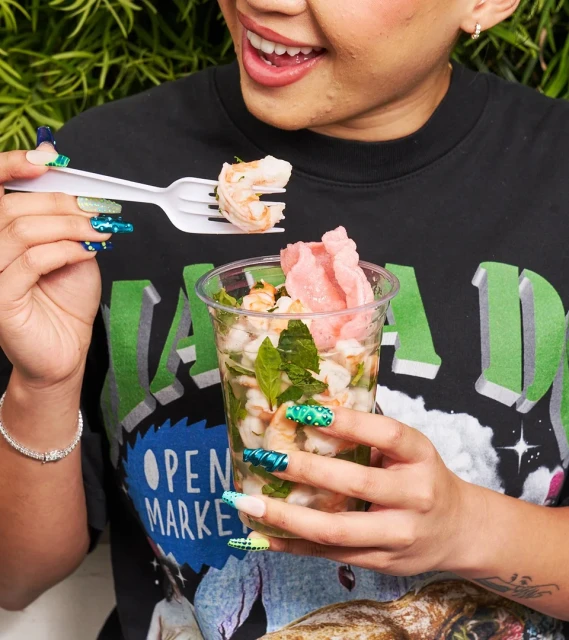 A person is eating shrimp ceviche from a clear plastic cup outdoors, showcasing fresh seafood and vibrant green foliage.