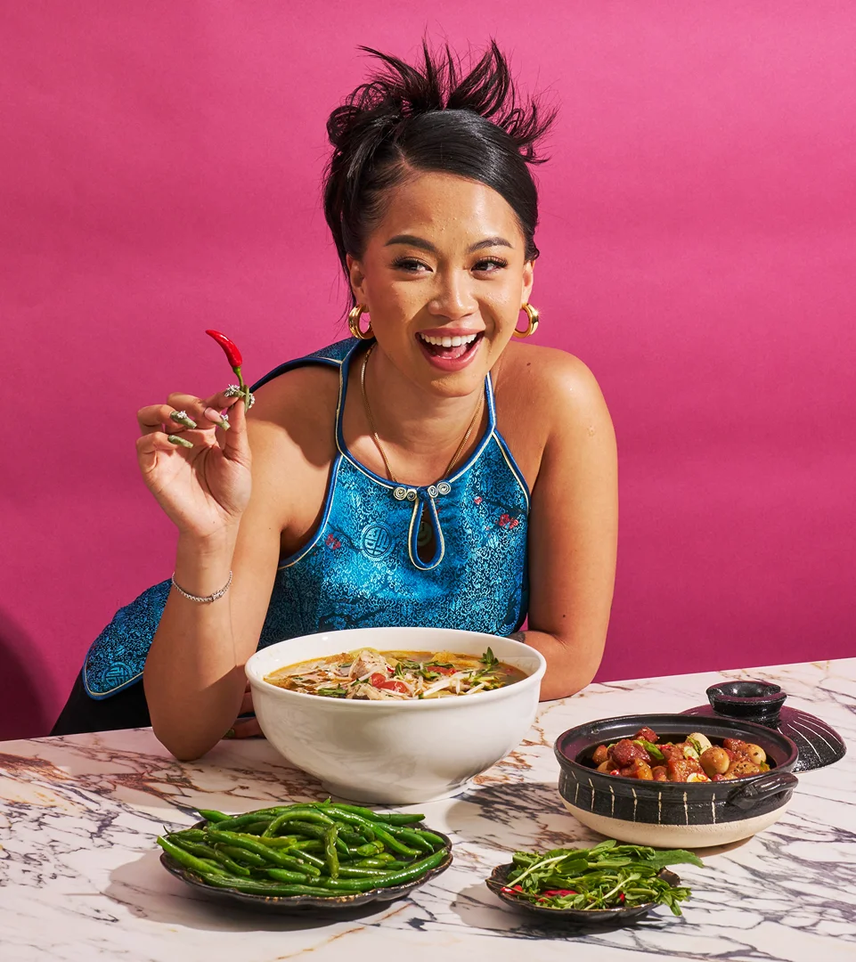 Smiling Asian woman in teal top holds a red chilli pepper next to bowls of food on a marble table against a pink background.