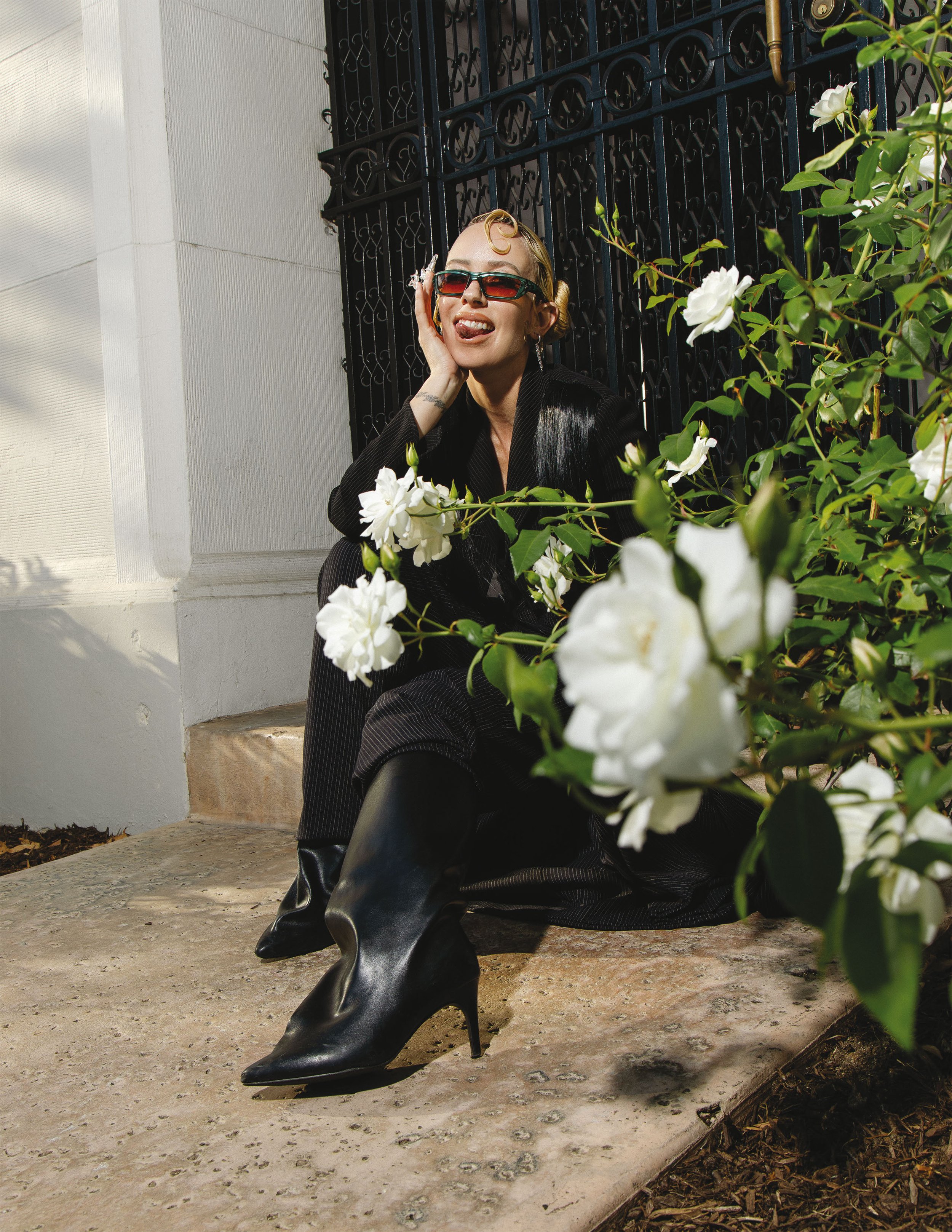 Blonde adult woman sitting outside on a concrete step wearing black clothing, boots and sunglasses, with a white rose bush beside her.