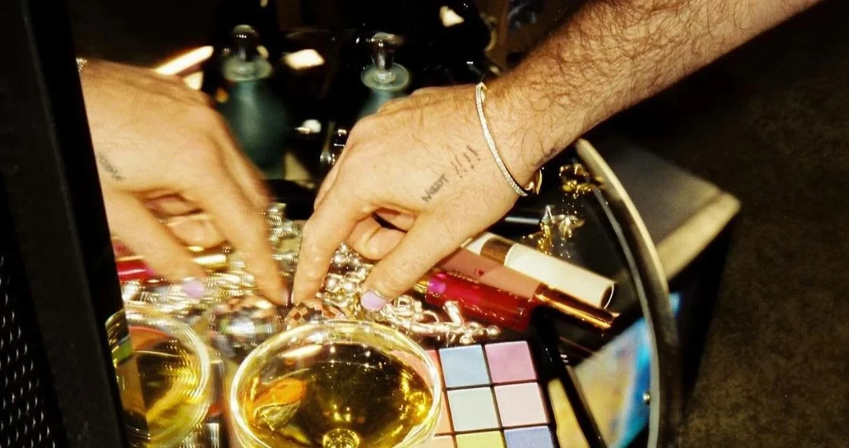 Close-up shot of a male's hand adorned with a diamond bracelet, resting near jewelry, glasses and a make-up palette on a black surface.