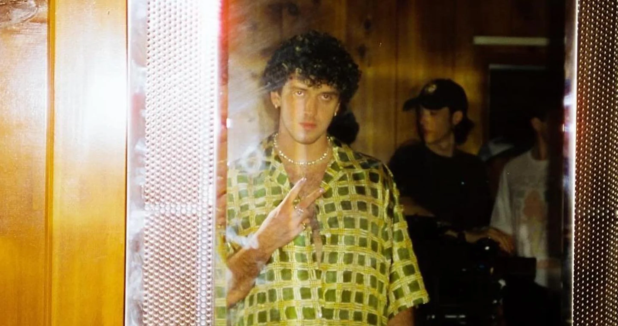Portrait of curly-haired man wearing patterned shirt, jewelry, gesturing peace sign indoors, with other people in background, low lighting.