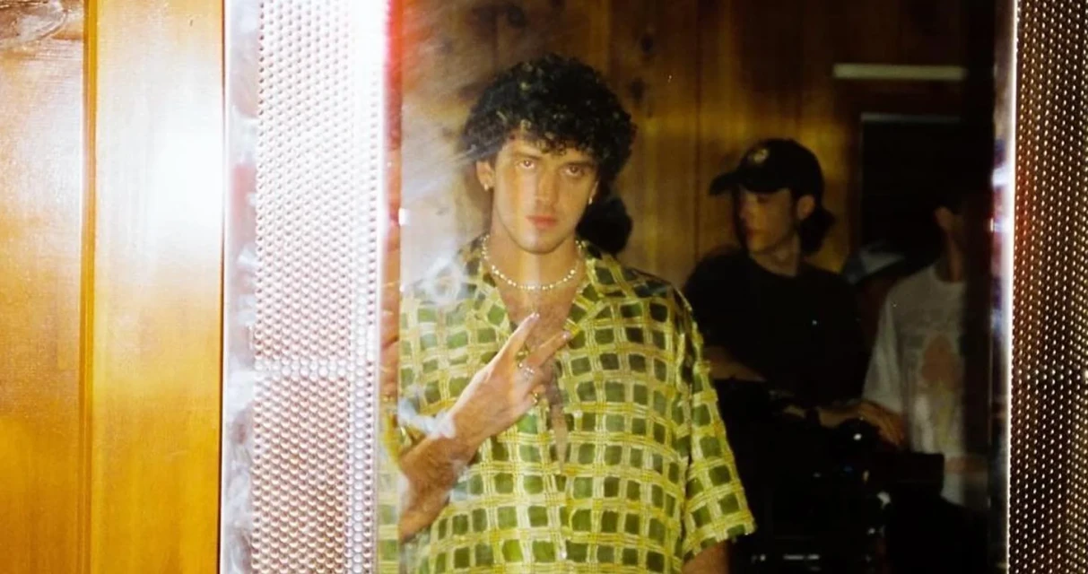 Portrait of curly-haired man wearing patterned shirt, jewelry, gesturing peace sign indoors, with other people in background, low lighting.