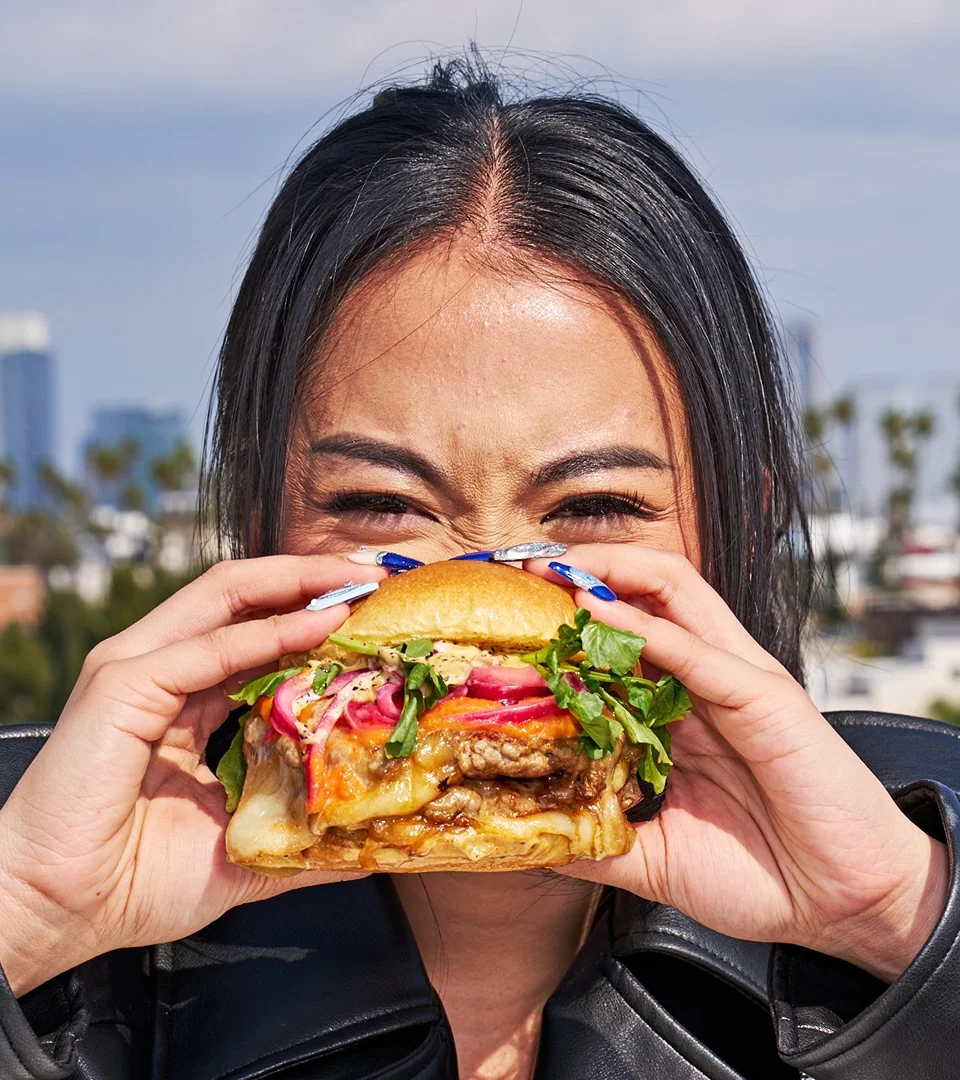 Adult female smiles while holding a loaded burger with toppings; outdoor urban setting with building silhouettes in background.