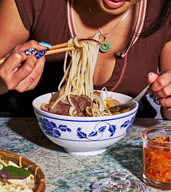 A person eats noodle soup with chopsticks and a spoon, showcasing a white and blue patterned bowl filled with delicious Asian cuisine.
