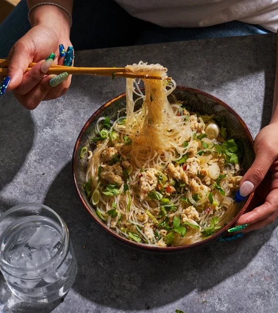 Overhead shot of hands using chopsticks to lift noodles from a bowl, next to a glass of ice water on a grey surface.
