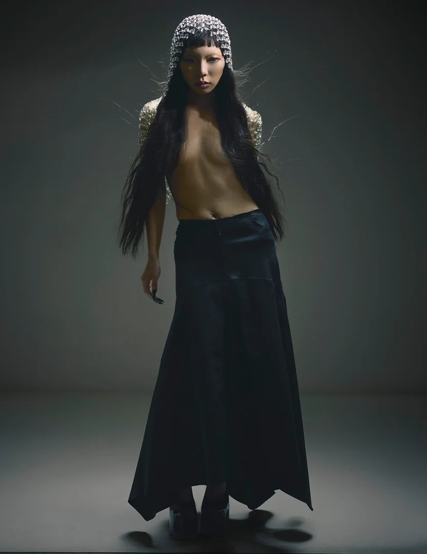 Fashionable young adult poses in dark skirt, beaded top and head covering in a studio setting, creating a striking visual statement.