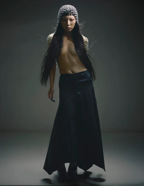 Fashionable young adult poses in dark skirt, beaded top and head covering in a studio setting, creating a striking visual statement.