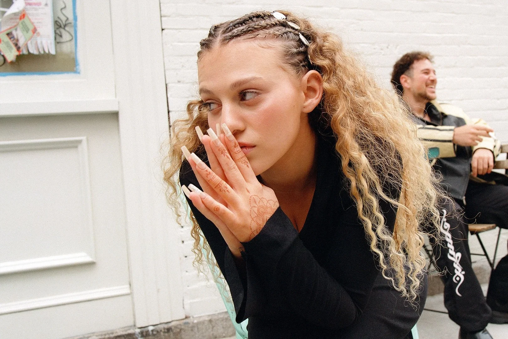 Portrait of woman with braided hair and long nails, posing with her hands up to her face, and a man sitting behind her in the background.