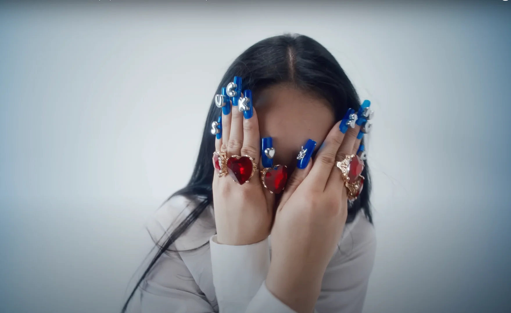Portrait of a woman with long dark hair wearing a white shirt, covering part of her face with hands adorned with large rings and blue nails.
