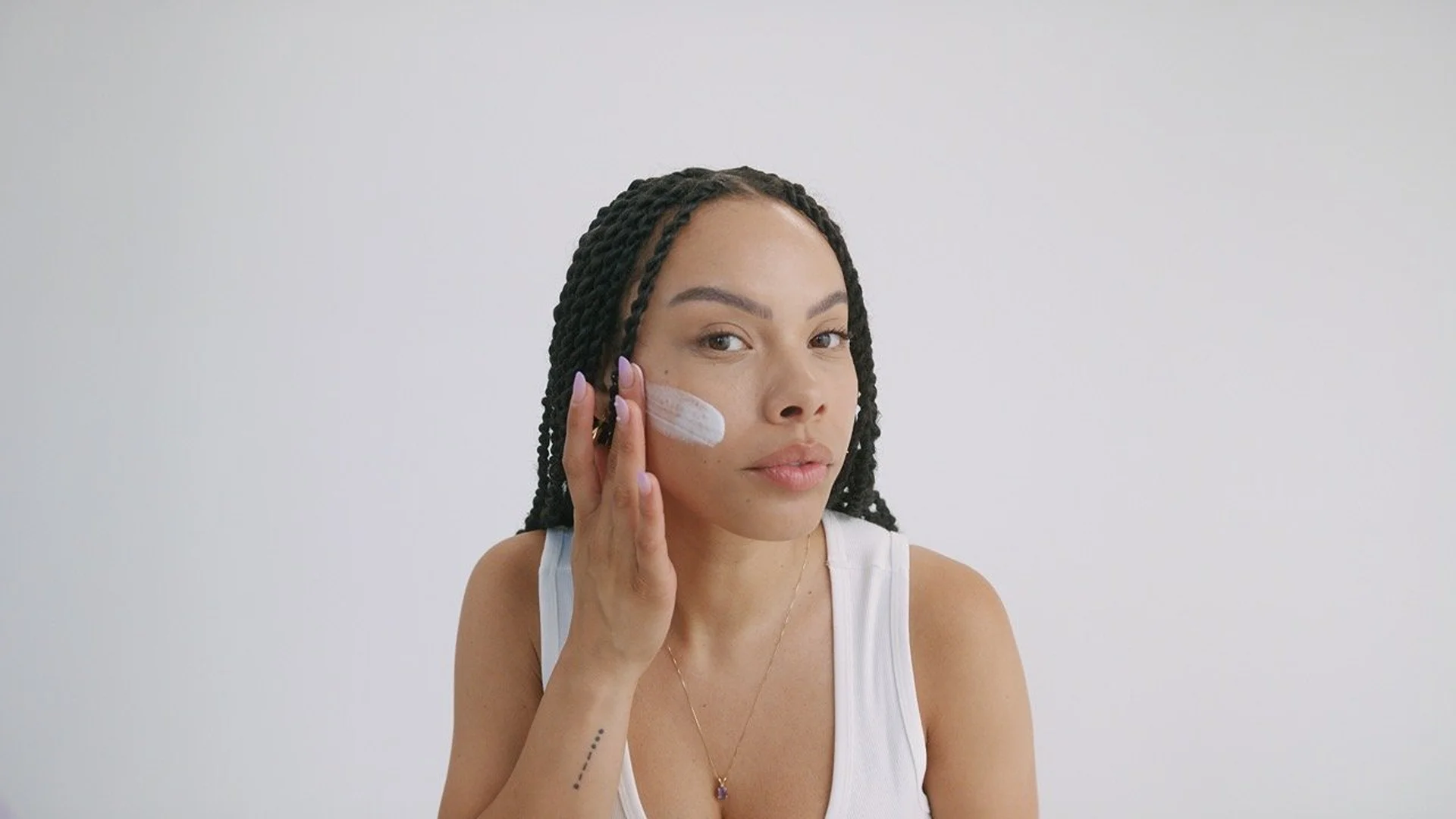 A woman with box braids applies white cream to her face in a close-up shot against a plain white background, showcasing skincare routine.