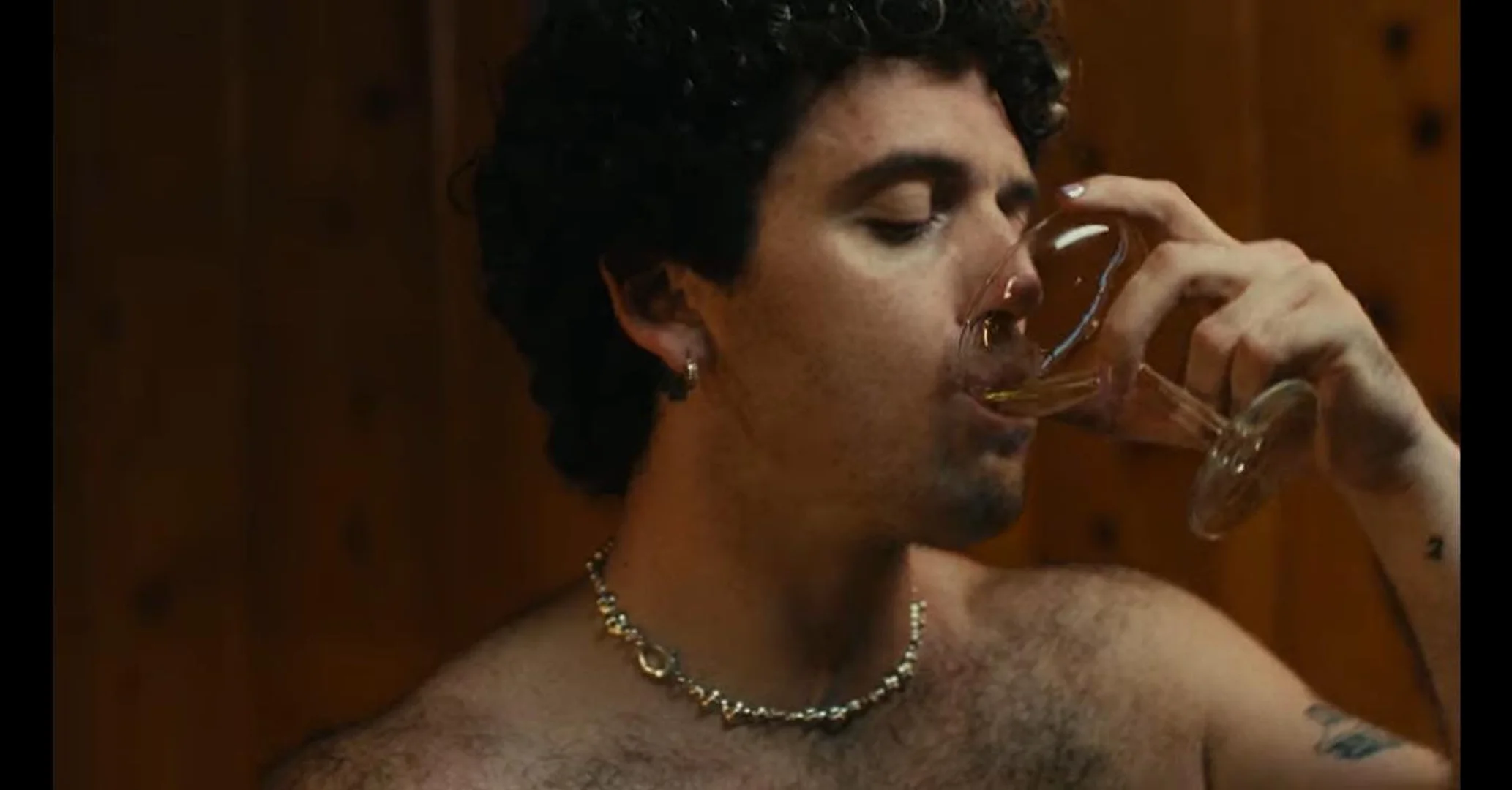 Close-up portrait of a man with dark curly hair drinking from a clear wine glass against a wooden paneled wall
