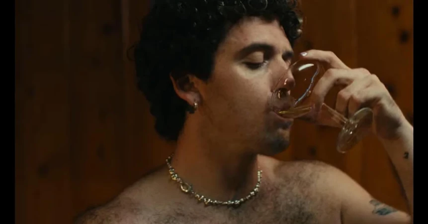 Close-up portrait of a man with dark curly hair drinking from a clear wine glass against a wooden paneled wall