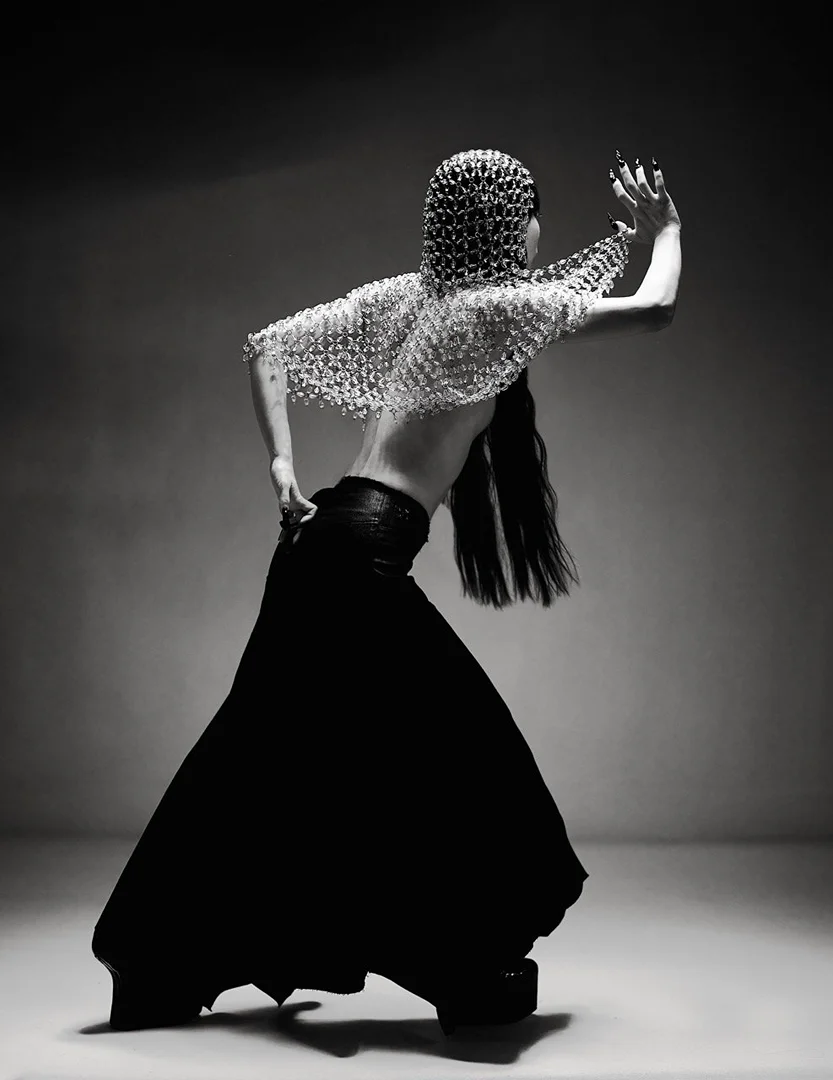 Woman in avant-garde fashion strikes a pose against a grey background. She's wearing a beaded headpiece and a long black skirt.