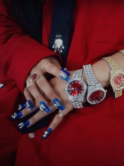 Close-up of manicured hands adorned with diamond rings, wristwatches, and a red blazer sleeve, emphasizing luxury and fashion.