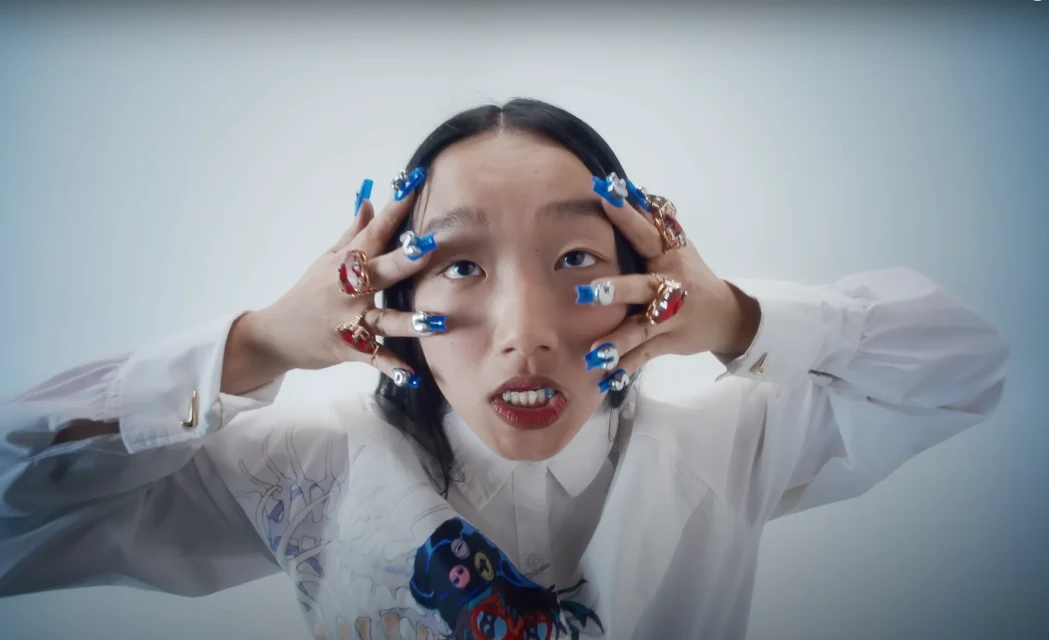 Woman with artistic blue nails, red rings, and white shirt poses in a studio with light blue background, looking directly at the camera.