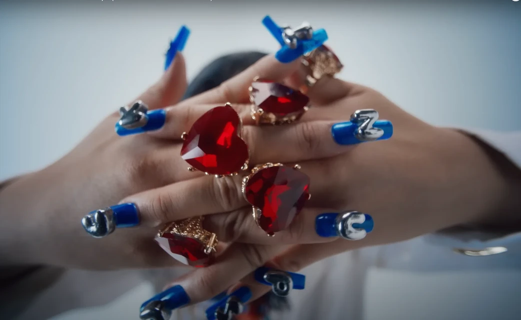 Close-up of two hands adorned with blue acrylic nails, silver letter charms, and heart-shaped ruby rings against a plain background.
