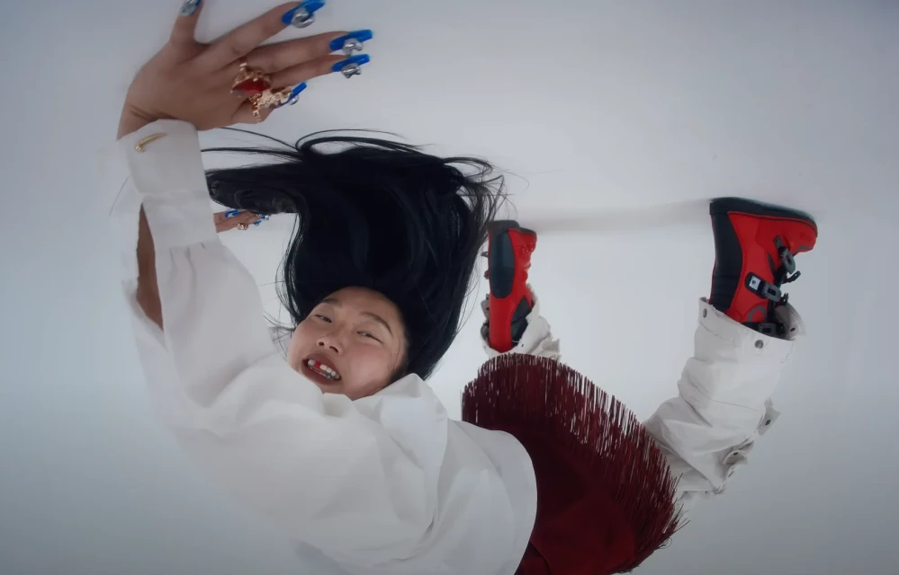Woman lying on a white surface wearing a white top, red skirt with fringe, red snow boots, and long blue painted nails with rings.