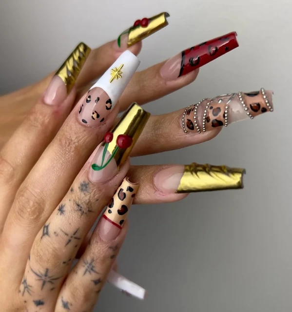 Close-up of hand with long, decorated nails, featuring animal prints, gold foil, and cherry designs, set against a plain background.