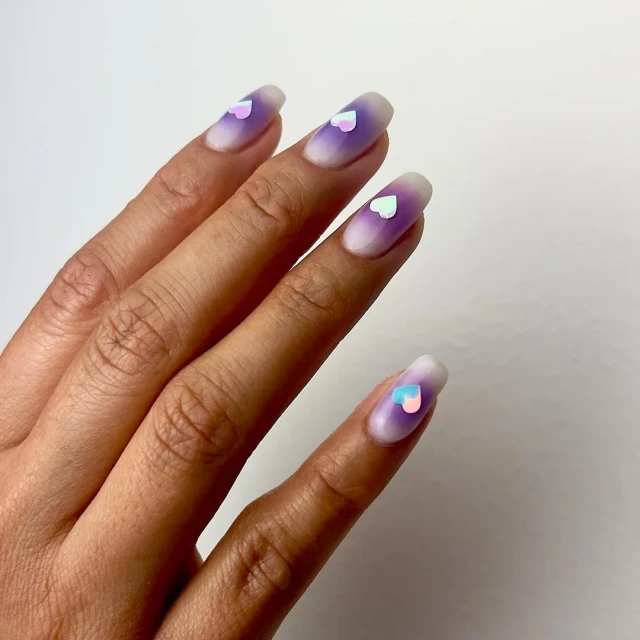 Close-up of a woman's hand with a gradient purple manicure and heart-shaped nail art, set against a bright white background.