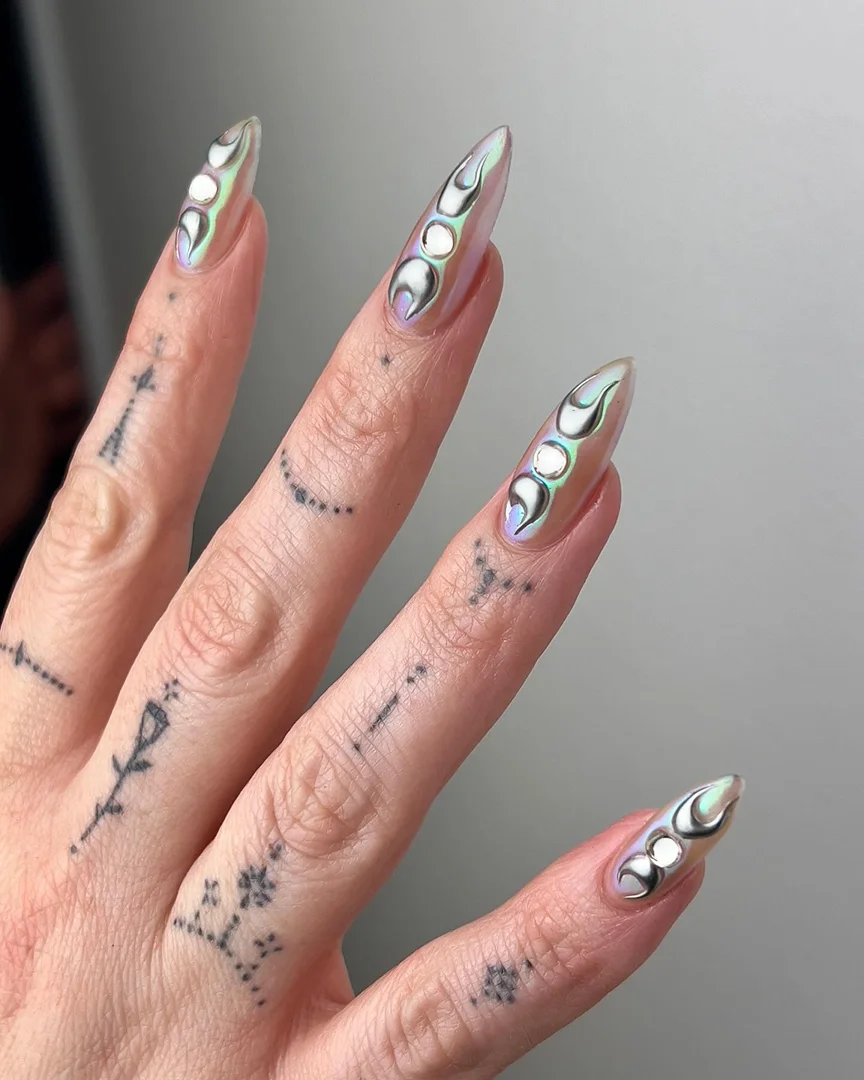Close-up of a woman's hand with long, pointed nails featuring a white moon phase design, along with small tattoos on the fingers and hand.