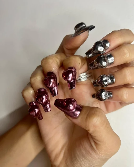 Close-up of a person's hands with silver and purple heart-shaped nail art, showcasing unique and reflective nail designs against white background.
