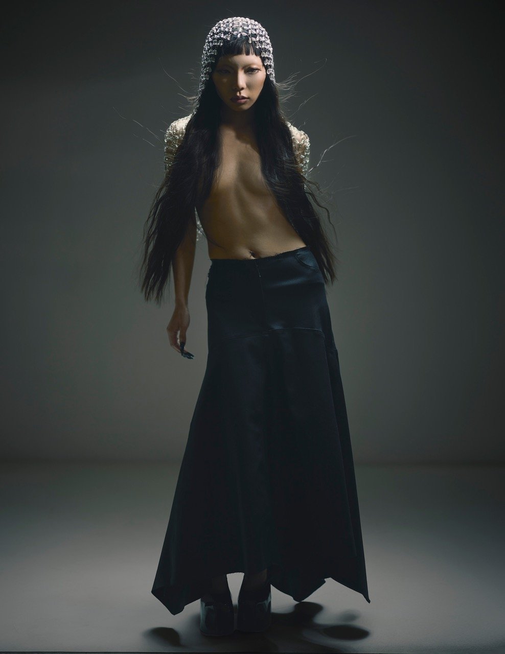 Adult female fashion model posing in black skirt and beaded head covering in photo studio with dark background and soft lighting.
